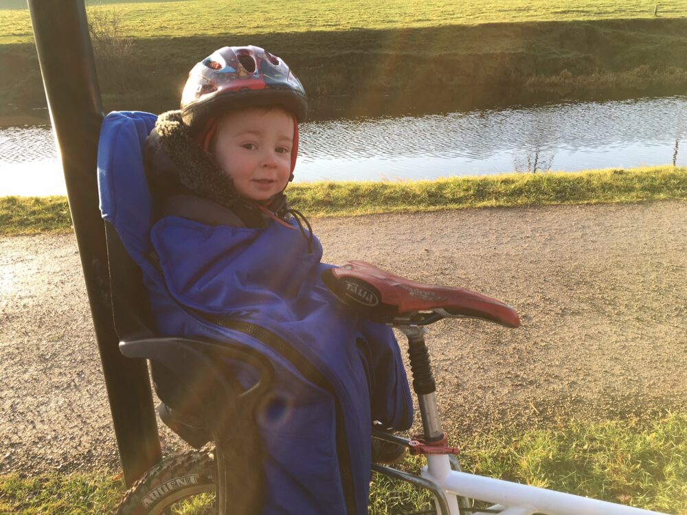 A young toddler wrapped up to keep warm and wearing a bike helmet, strapped into a rear-mounted bike seat