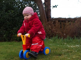 Image shows a two year old toddler on a small balance bike riding down a slight grass slope in their garden