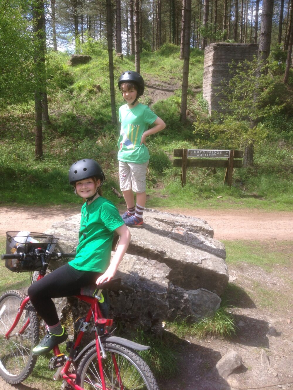 Forest of Dean Family Cycling Route A girl and boy resting next to a big rock in the forest, wearing bike helmets and smiling at the camera
