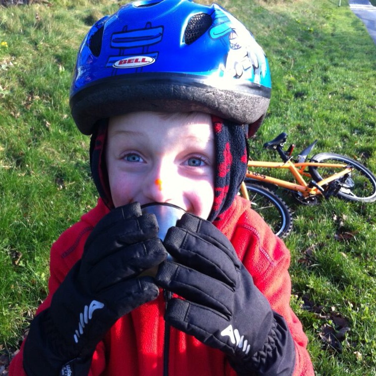 Winter cycling with kids: A young boy wearing thick cycling gloves and a helmet, enjoying some hot soup from a flask