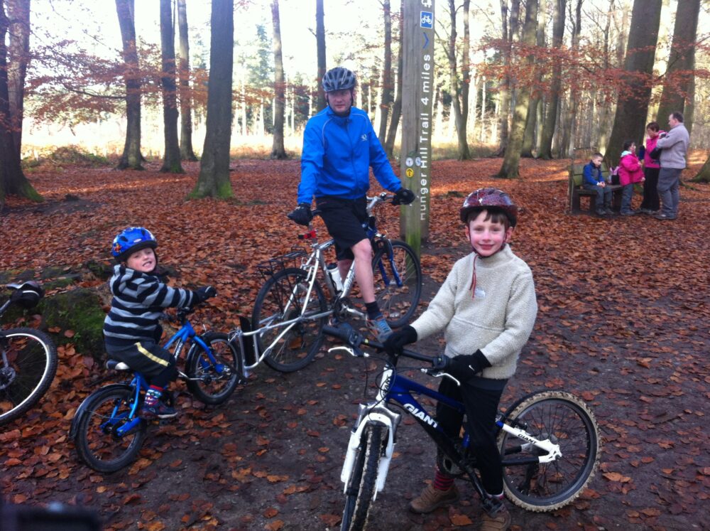 How to choose the right size kids' bike: A dad and two boys in the woods. One boy is on his own bike, the other is on a tagalong