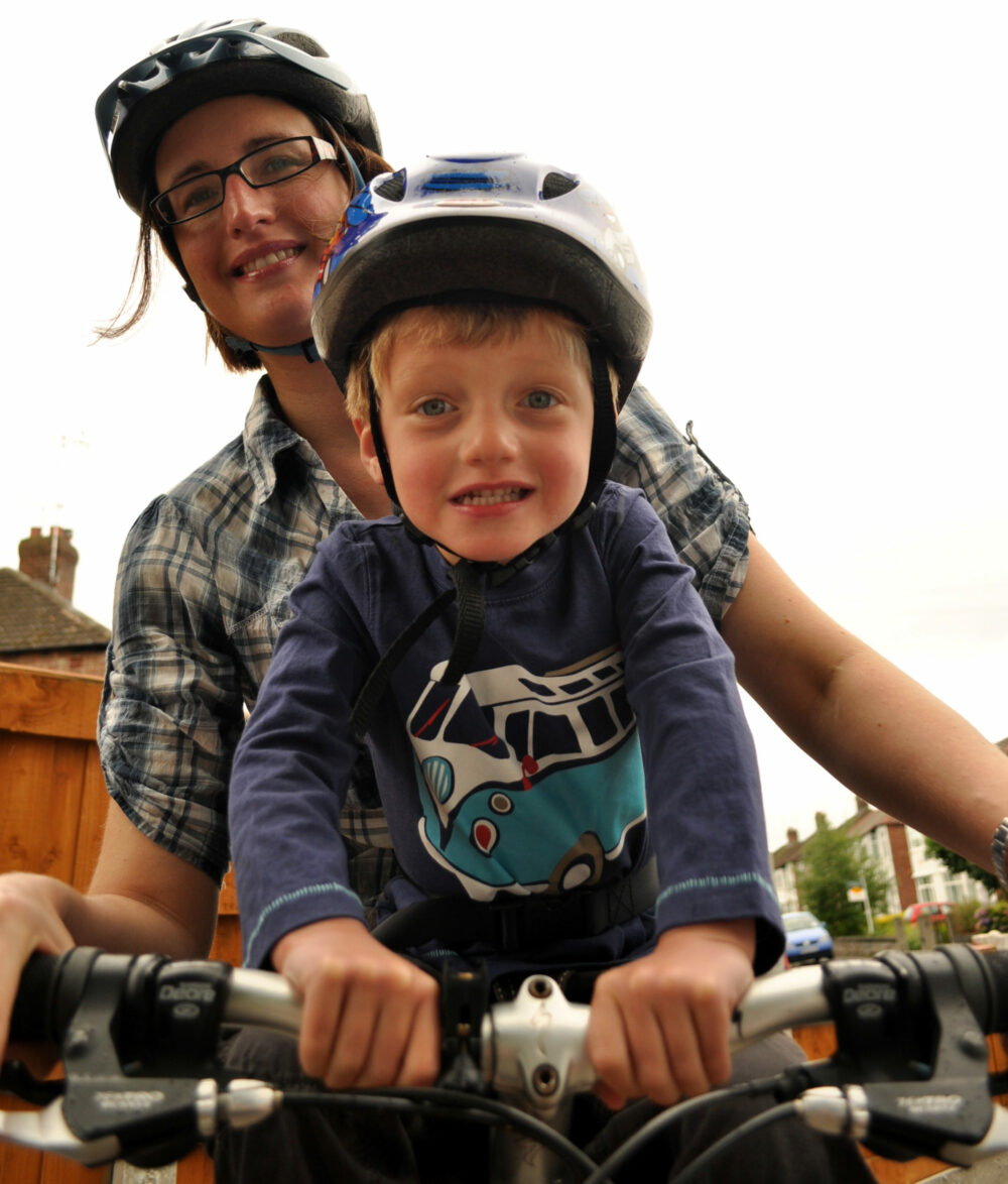 A close up of Karen and her son on a bike, he's sitting on a front-mounted bike seat and wearing a helmet, holding onto the handlebars. Karen is leaning round to smile at the camera