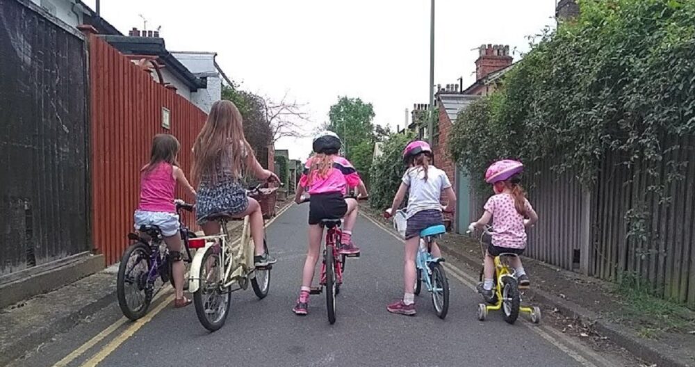 Syvlia Go – Girls on Bicycles Bikes for girls: a row of five little girls of different ages, mounted on bicycles, seen from behind