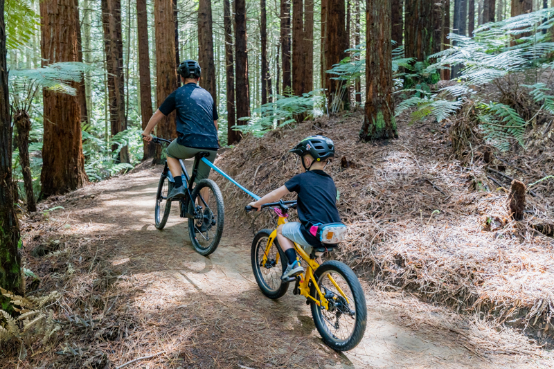 A cyclist climbing a hill With their child's bike tethered to theirs, with The Shotgun MTB Tow Rope