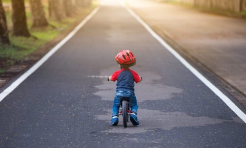 Best bike for a 1 year-old: a small boy on a pre-balance bike, seen from behind on a paved cycle path with trees lining the edges