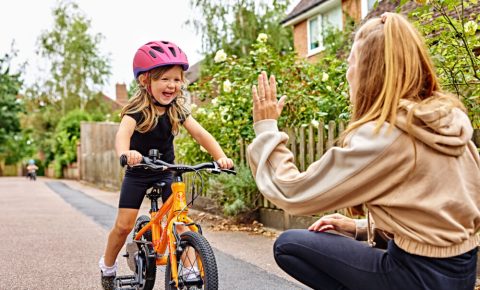 A little girl wearing a bike helmet riding a pedal bike towards her mum, who crouches and holds a hand out for a high five
