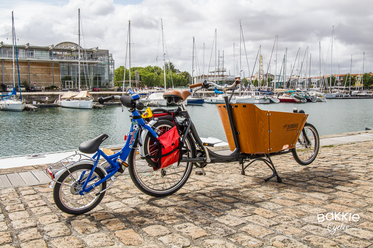 A child's bike hitched up to a Bakfiets, with the front wheel secured inside a Bakkie Cycles pannier bag