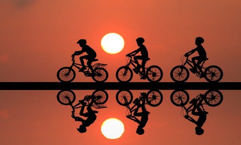 Black outline of children riding their bikes against the sunset, with mirror image in water underneath them