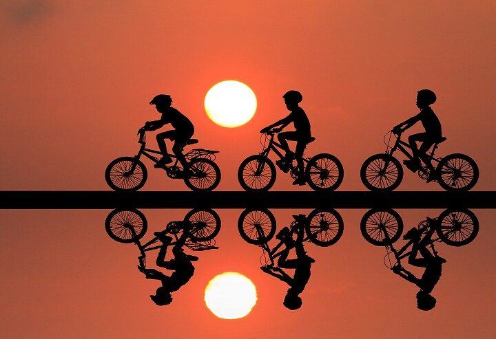Black outline of children riding their bikes against the sunset, with mirror image in water underneath them