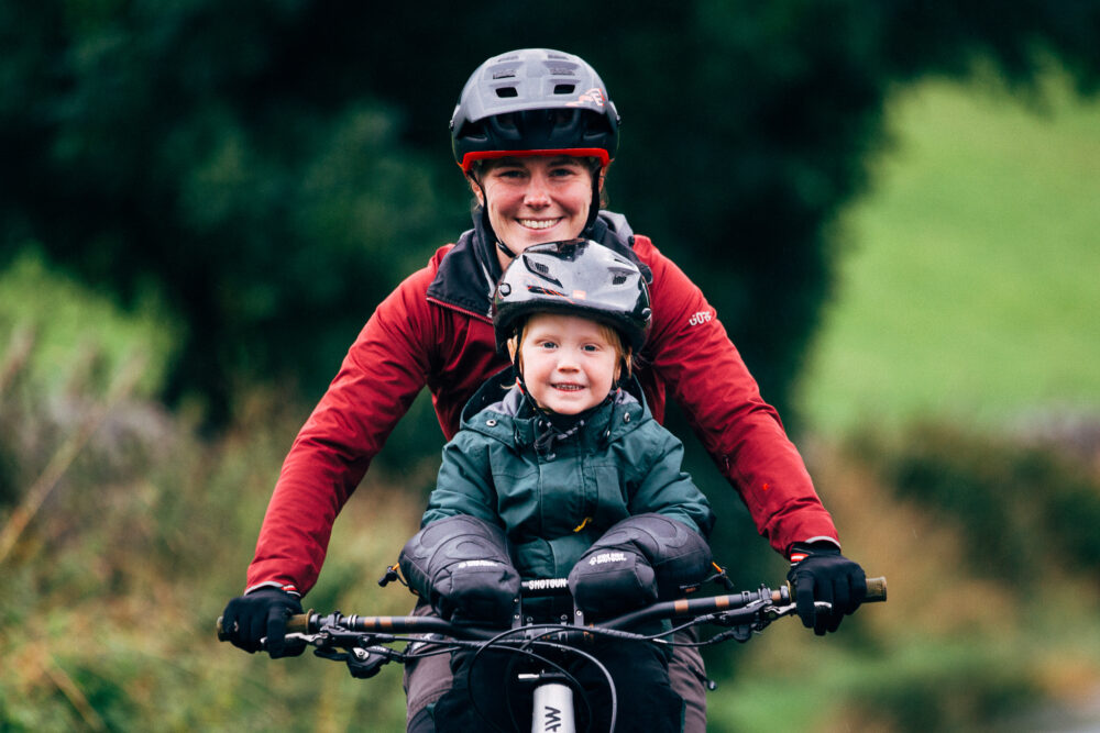 Small toddler on front bike seat keeping his little hands warm in the pogies. He has a big smile on his face, and is mum is behind him steering the bike,