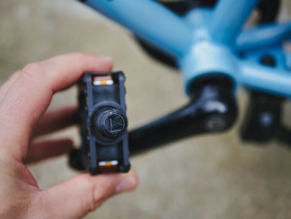 Photo of a hand holding a bike pedal with the letter L on it, with a blue child's bike in the background - demonstrating that there is a correct way to put the pedals back into a kids bike 