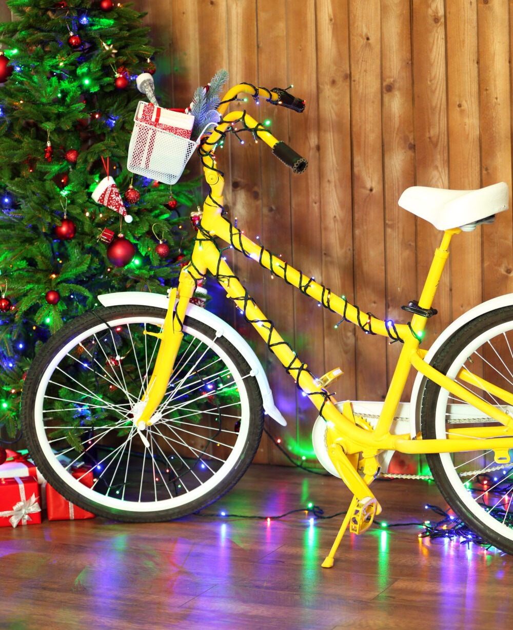 Child's bike with fairy lights wrapped around it near a Christmas tree with presents in the basket