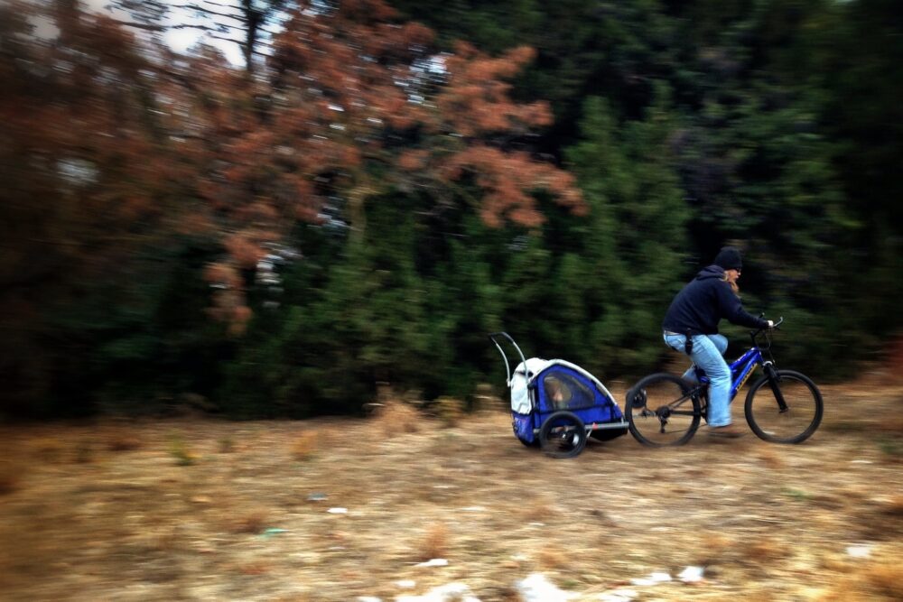 Photo of a woman cycling in the autumn pulling a bike trailer with a child in it behind her.  A cheap bike trailer can help give year round transport without the costs of a car, petrol or parking 