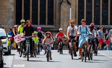 What is Kidical Mass? This photo shows a group of cylclists of all ages riding through a street in Bath, UK, on a Kidical Mass ride