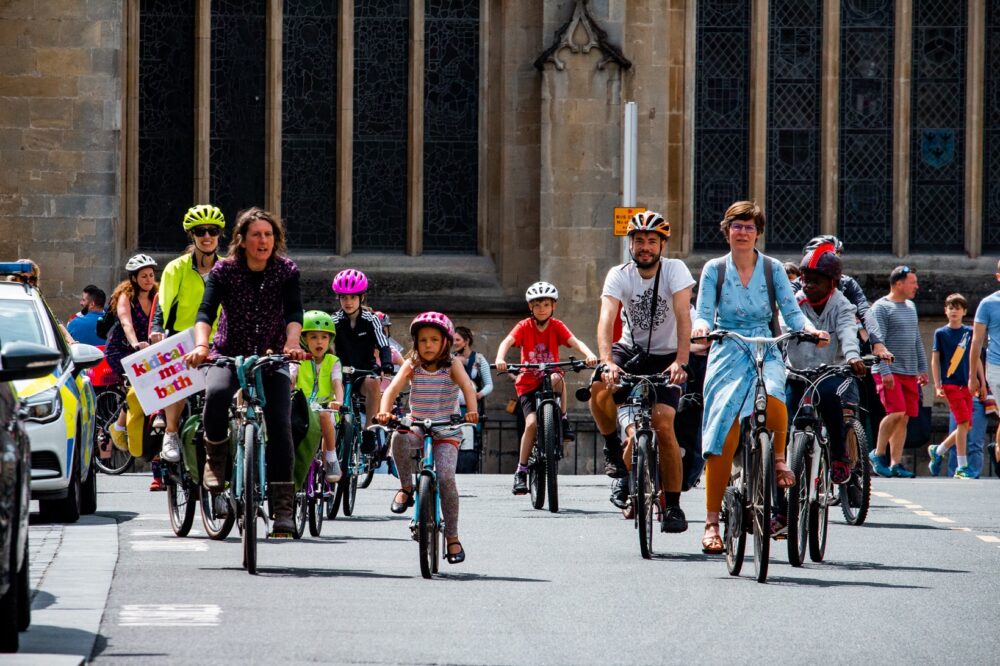 What is Kidical Mass? This photo shows a group of cylclists of all ages riding through a street in Bath, UK, on a Kidical Mass ride