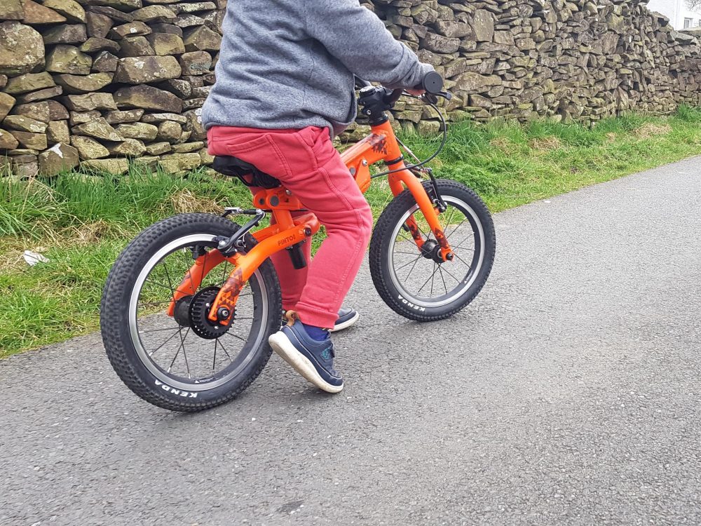 Photo of the Black Mountain PINTO being used in balance bike mode, as part of this detailed review. Shows body and legs of a small child riding an orange PINTO along a strip of tarmac, with a grass bank and stone wall in the background. The child is wearing red trousers and blue shoes and a grey jacket.