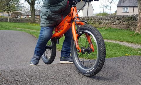 Black Mountain PINTO in balance bike mode. Photo of a young child riding an orange balance bike with Black Mountain written on the frame on a tarmac pump tack. Child is wearing blue jeans and blue shoes.