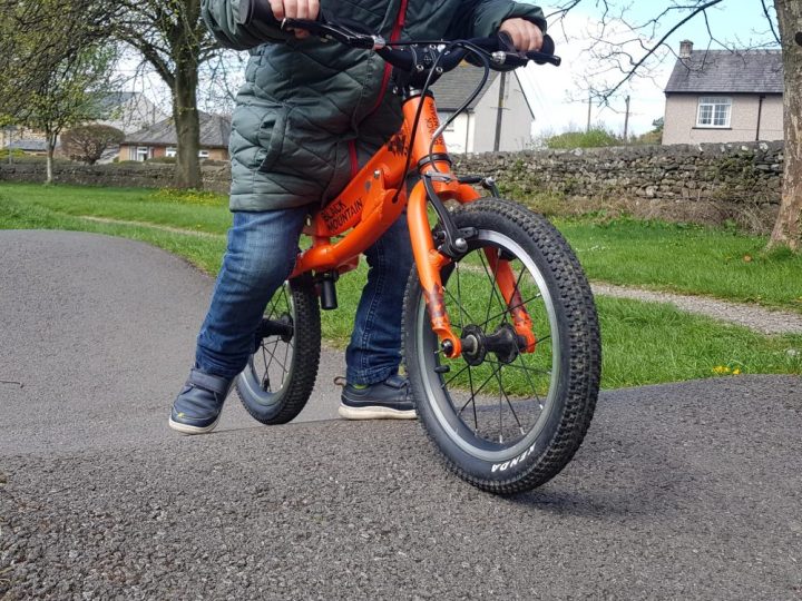 Black Mountain PINTO in balance bike mode. Photo of a young child riding an orange balance bike with Black Mountain written on the frame on a tarmac pump tack. Child is wearing blue jeans and blue shoes.