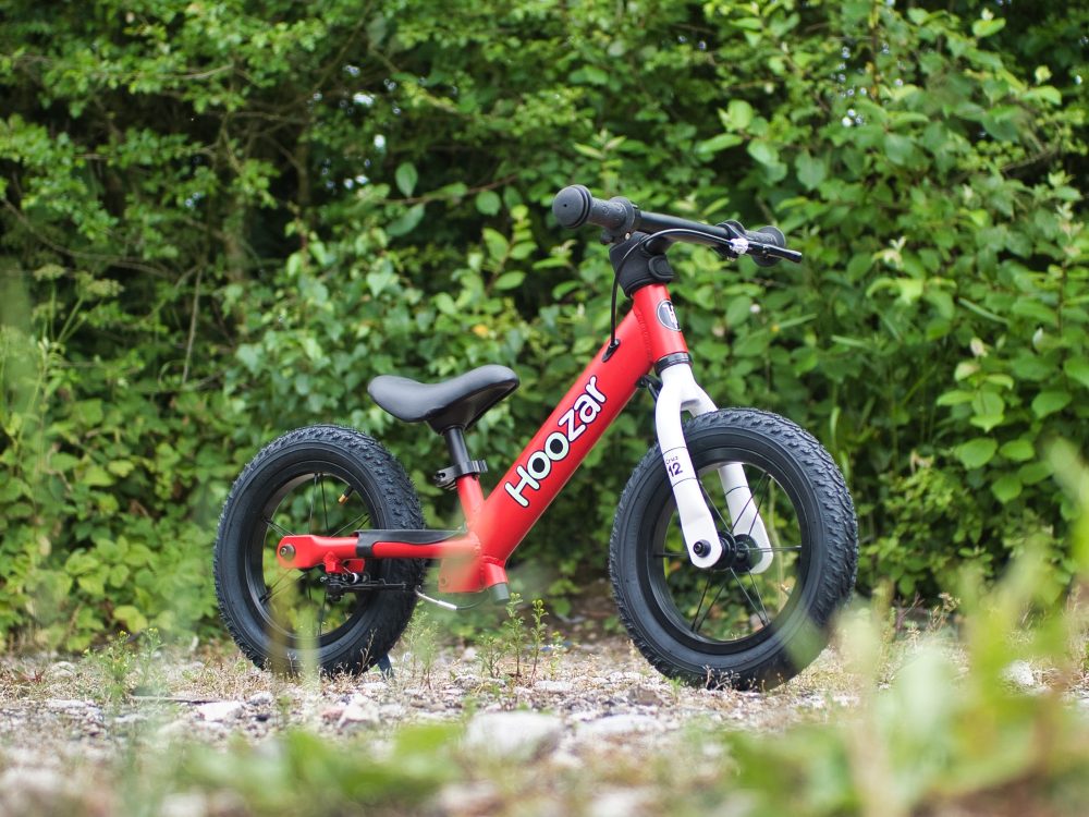 The Hoozar Cruz balance bike for children aged 2 and over - the photo shows the red balance bike we are reviewing, set against a backdrop of trees