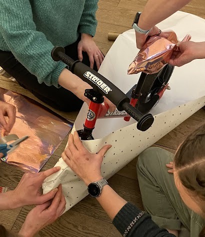Several adults seen from above, attempting to wrap a balance bike with wrapping paper