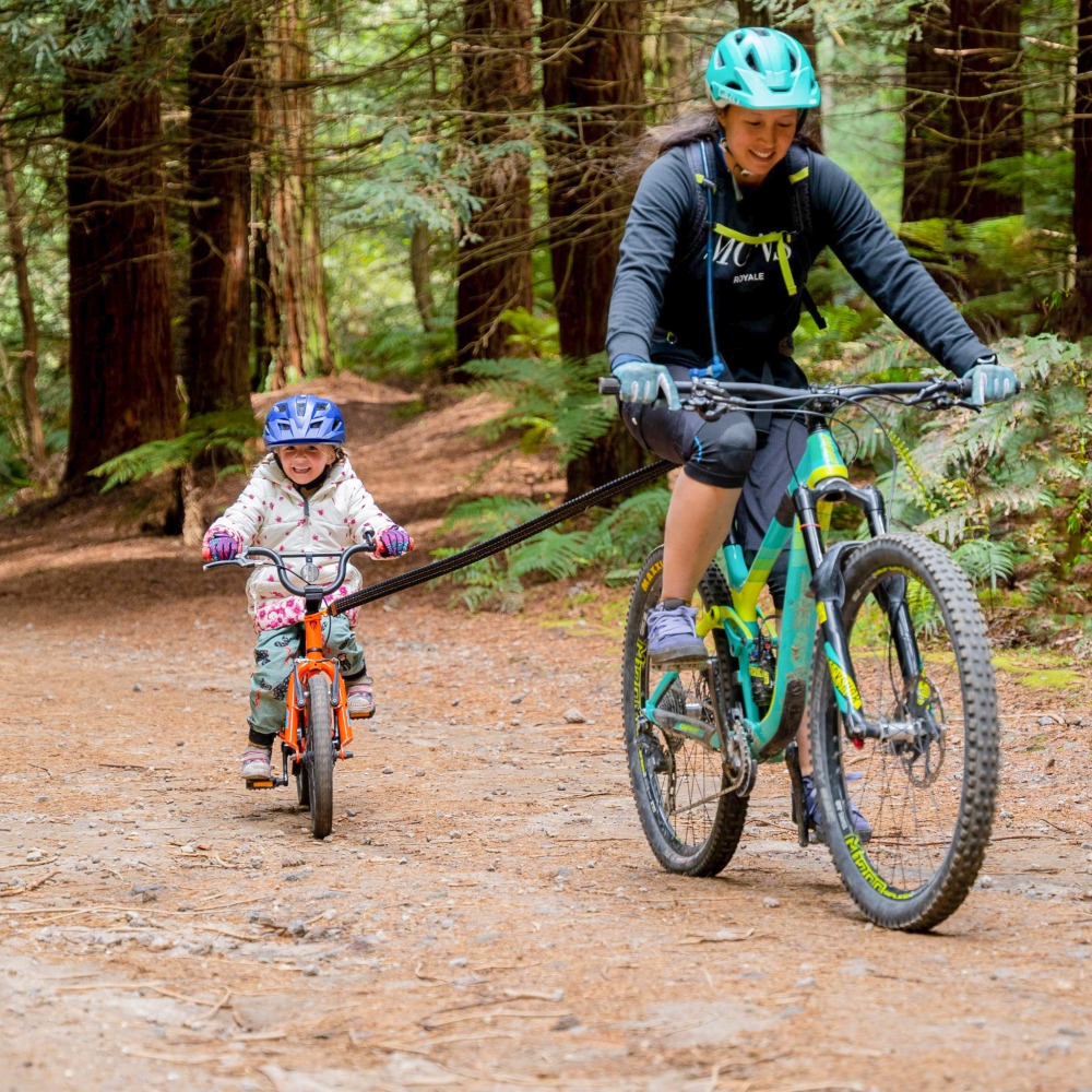 A mountain biking parent towing their child's bike with a tow rope, in the woods