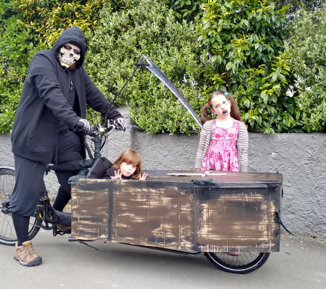 Halloween Costumes for Cycling Kids: An adult dressed as the grim reaper riding a cargo bike decorated as a coffin, with two children inside