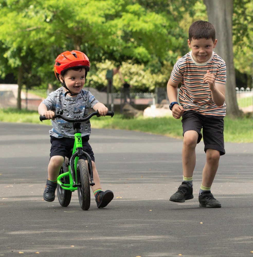 How to teach an autistic child to ride a bike: This photo shows a young boy riding a Kidvelo balance bike, and his older brother running alongside him, in a quiet street full of trees.