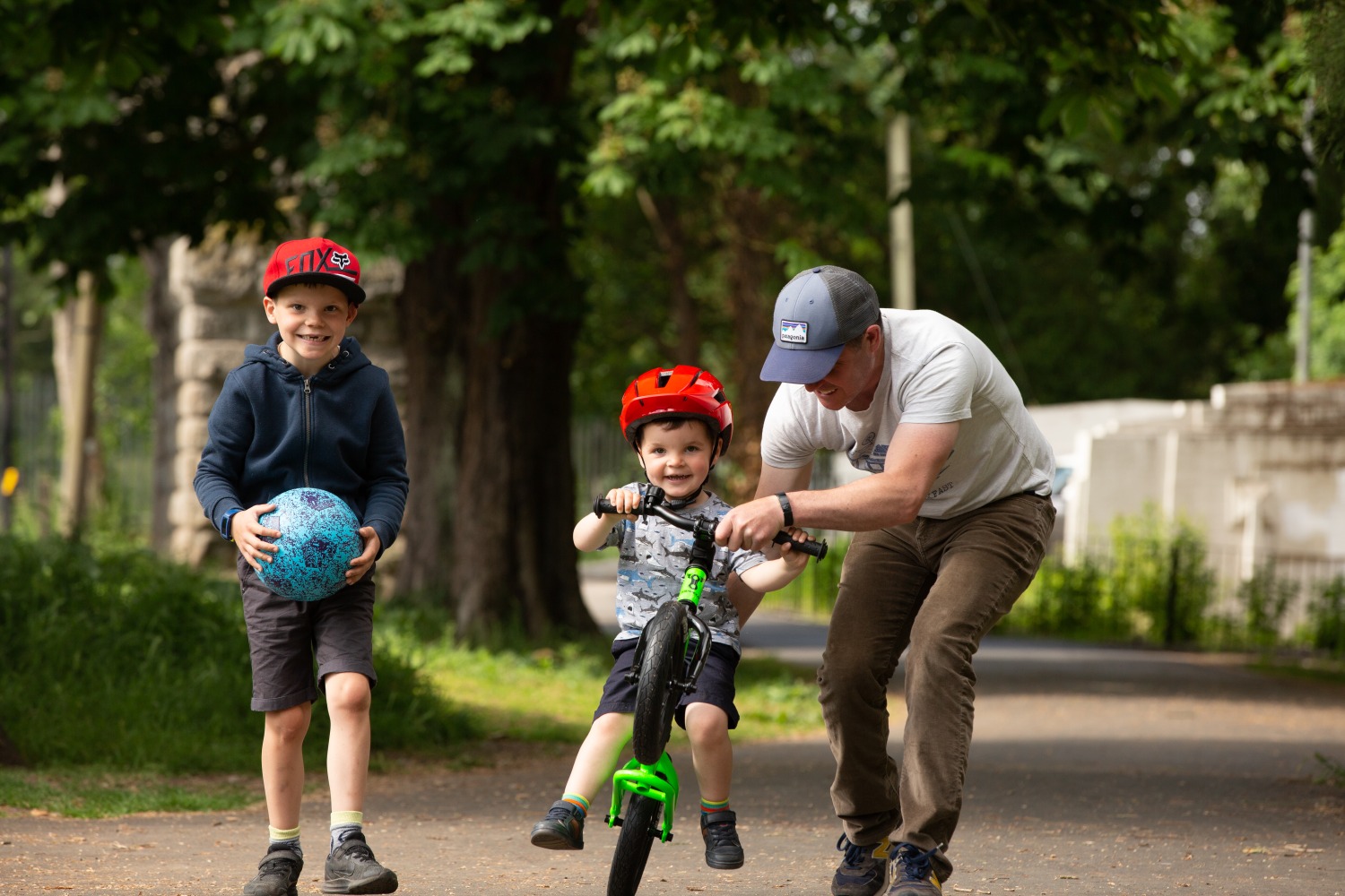 How to teach an autistic child to ride a bike: A young boy on a Kidvelo balance bike on a leafy suburb street. His dad is lifting the front of the bike to simulate a wheelie, and his brother is walking alongside him and holding a football