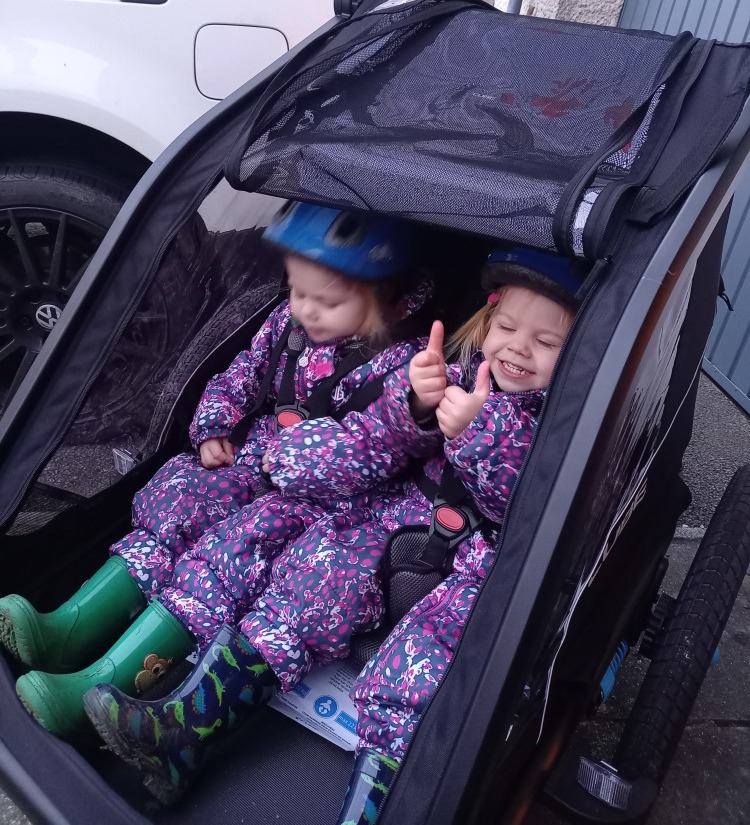 Two twin girls smiling and doing a thumbs up in a bike trailer