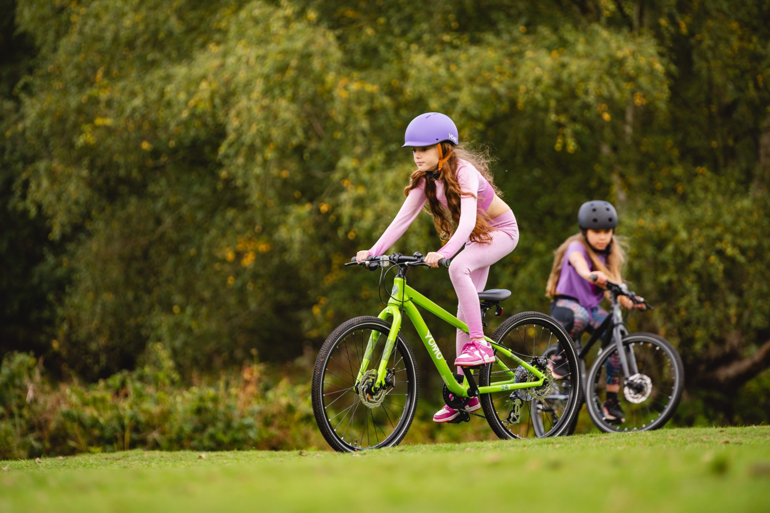 YOMO Kids bikes in action - two of the larger geared bikes being ridden by girls across some grass