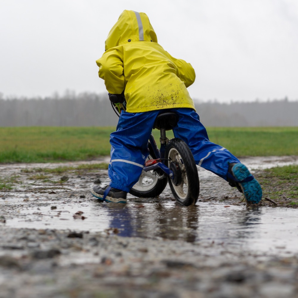 A boy in a raincoat and waterproof trousers rides his balance bike through a puddle