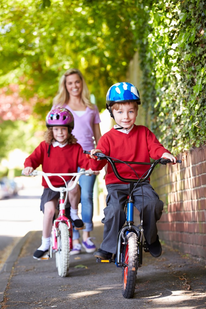 A mum walking along the pavement behind her two children who are cycling in school uniforms