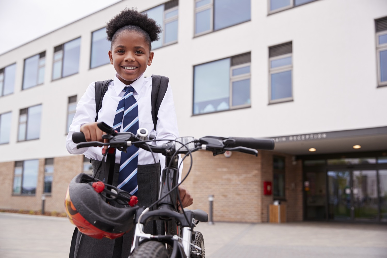 A girl in a school uniform, standing next to her bike, smiling, with a helmet dangling from the handlebar