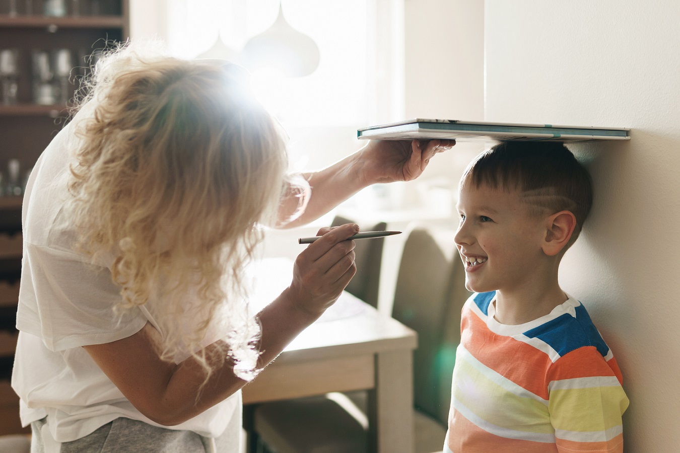 A woman holds a book above her son's head as she measures his height against a wall