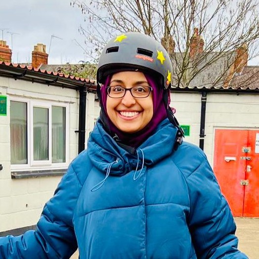Cycling in Ramadan - A headshot of Sarah Jassat, wearing a hijab under her bike helmet, standing in front of a building and smiling at the camera