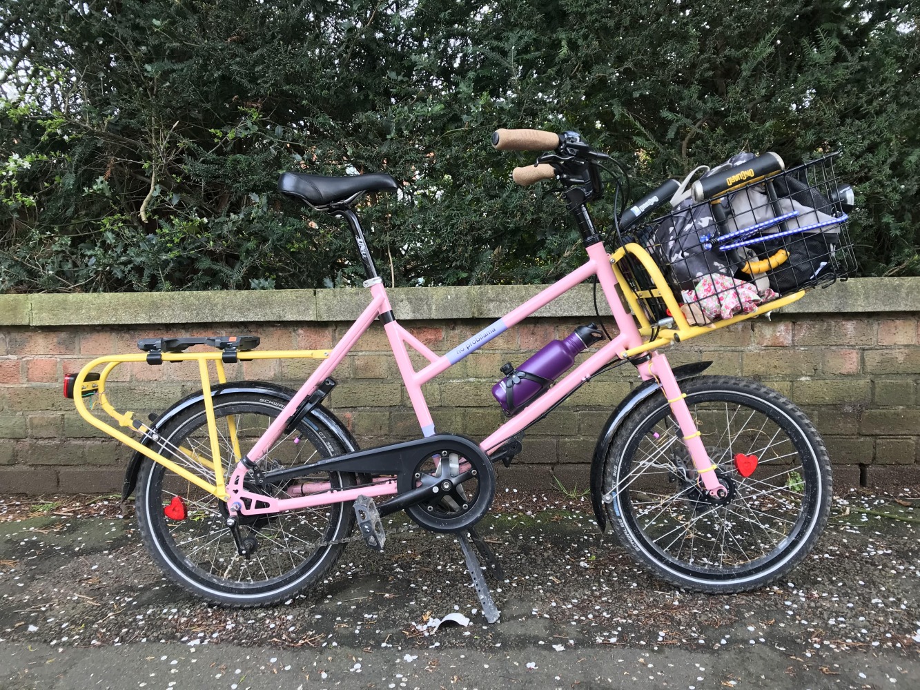 Cycling in Ramadan - A pink cargo bike with yellow front and rear racks, and a basket loaded up with luggage, parked in front of a wall. There's a bottle in the bottle cage.