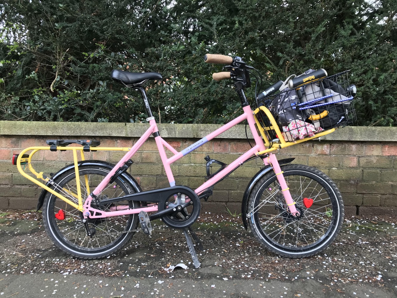 Cycling in Ramadan - A pink cargo bike with yellow front and rear racks, and a basket loaded up with luggage, parked in front of a wall. The bottle is now missing.