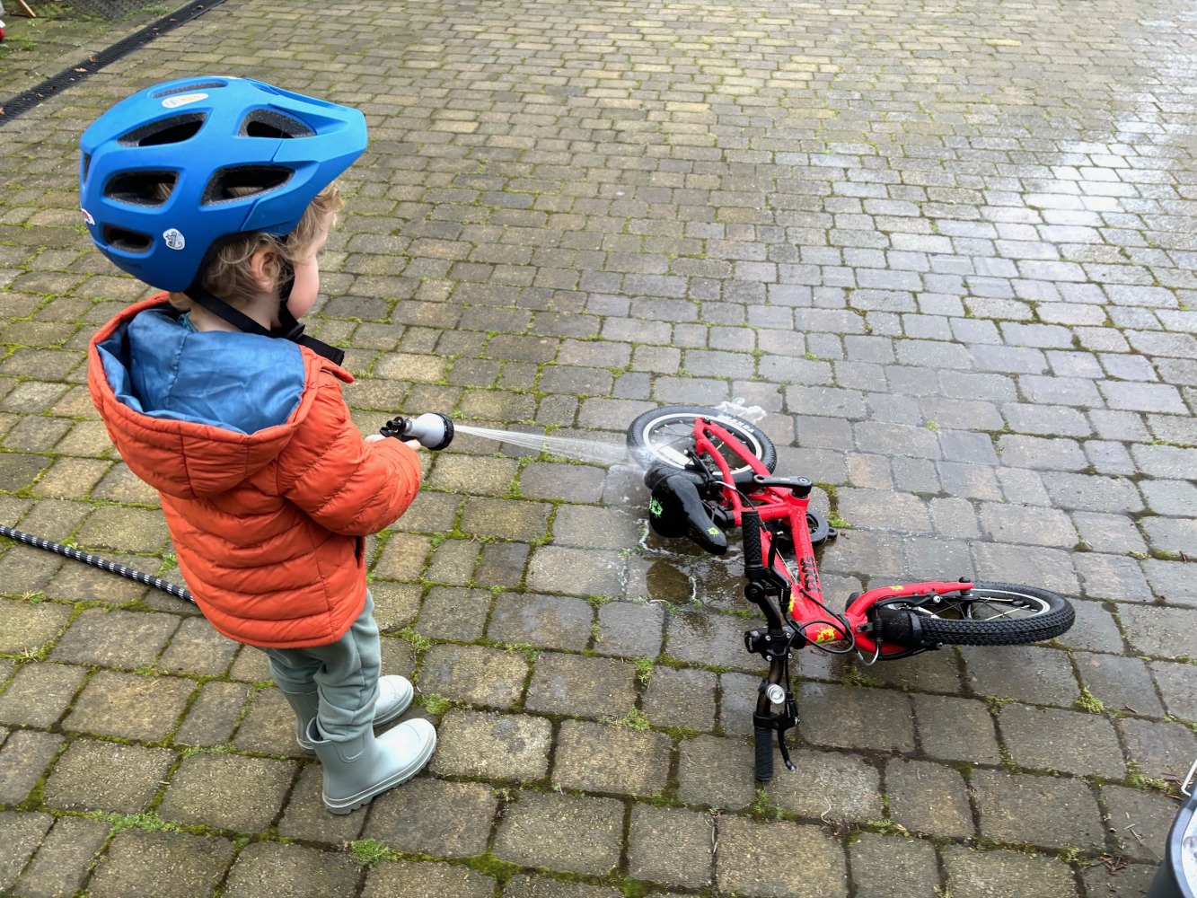 Boy in a orange jacket and blue helmet cleaning his bike with a hose