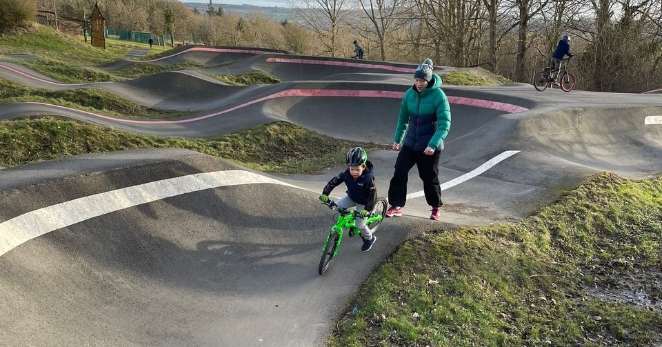 boy on a green bike riding a pump track with an adult behind him giving a push