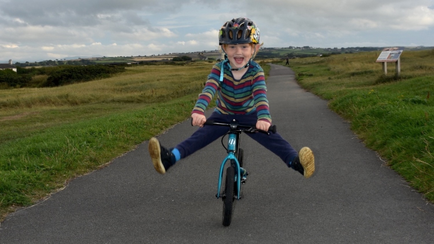 A toddler riding a balance bike towards the camera, smiling and lifting her legs out wide