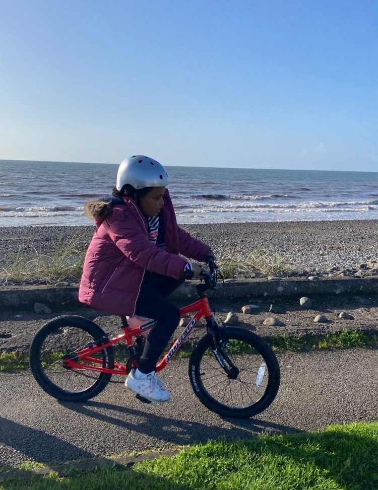 A girl in a pink coat and silver helmet riding the kidvelo rookie 18 along the sea front