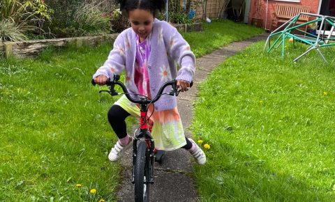 A girl in a pink cardigan and bunchies riding the kidvelo rookie 18 in her garden