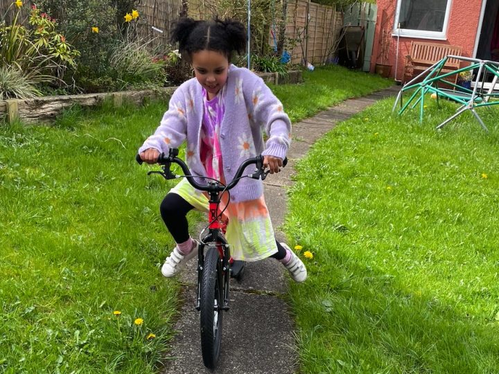 A girl in a pink cardigan and bunchies riding the kidvelo rookie 18 in her garden