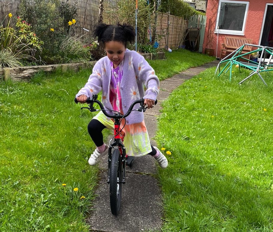 A black girl in a pink cardigan and bunchies riding the kidvelo rookie 18 in her garden