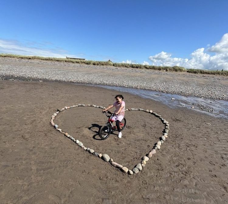 a young girl with afro hair standing within a heart made of rocks on a beach on her kidvelo rookie 18 bike