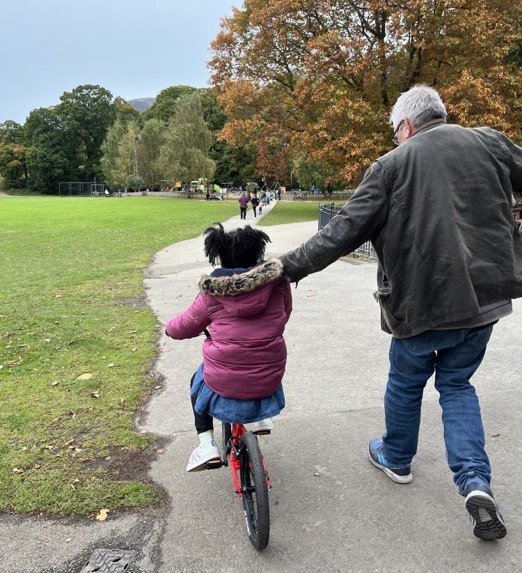 A black girl in a pink coat and afro hair riding the kidvelo rookie 18 in the park with her grandad holding on to her to keep her steady