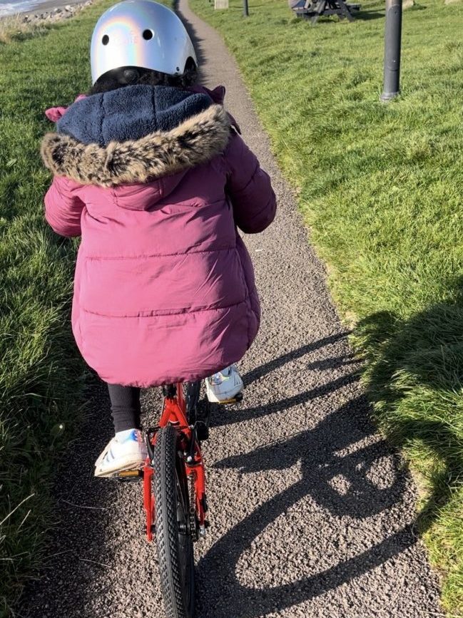 A girl in a pink coat and silver helmet riding the kidvelo rookie 18 along the sea front