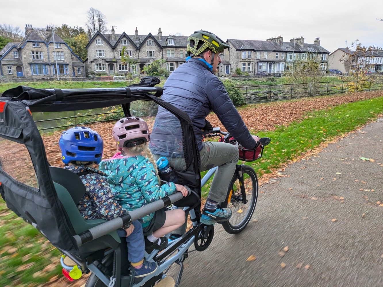 Decathlon elops longtail cargo bike review campaign for cycling infrastructure: a family cycling by the river on a winter day along a river, the two children are wearing colourful helmets and clothes and are looking away from the camera while their dad cycles