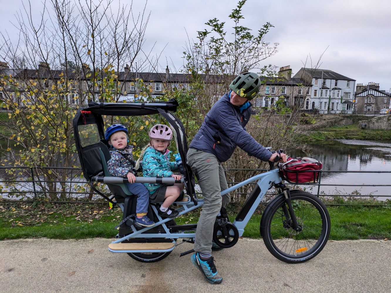 a family cycling by the river on a winter day along a river, the two children are wearing colourful helmets and clothes and are looking at the camera while their dad cycles