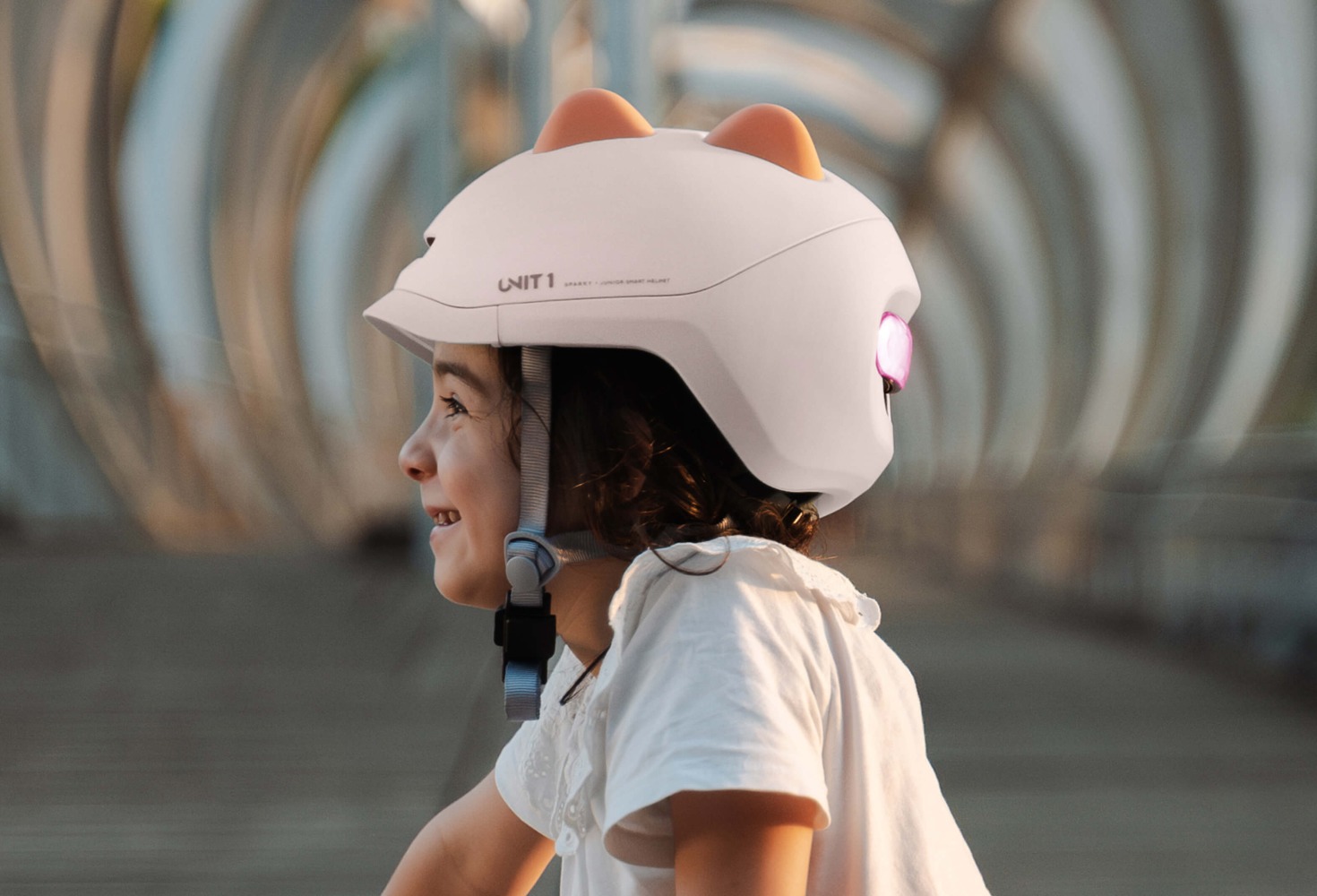 A little girl seen from the side, smiling an swearing a pink Sparky helmet with silicone dinosaur spikes on top and a rear light